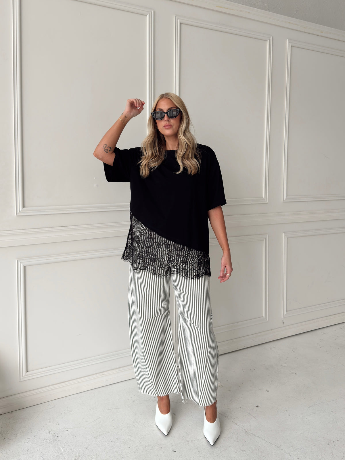 A woman modeling a black short-sleeve top with a sheer black lace ruffle hem, paired with white and olive vertically striped wide-leg pants and white pointed-toe heels, standing in front of a white paneled wall.