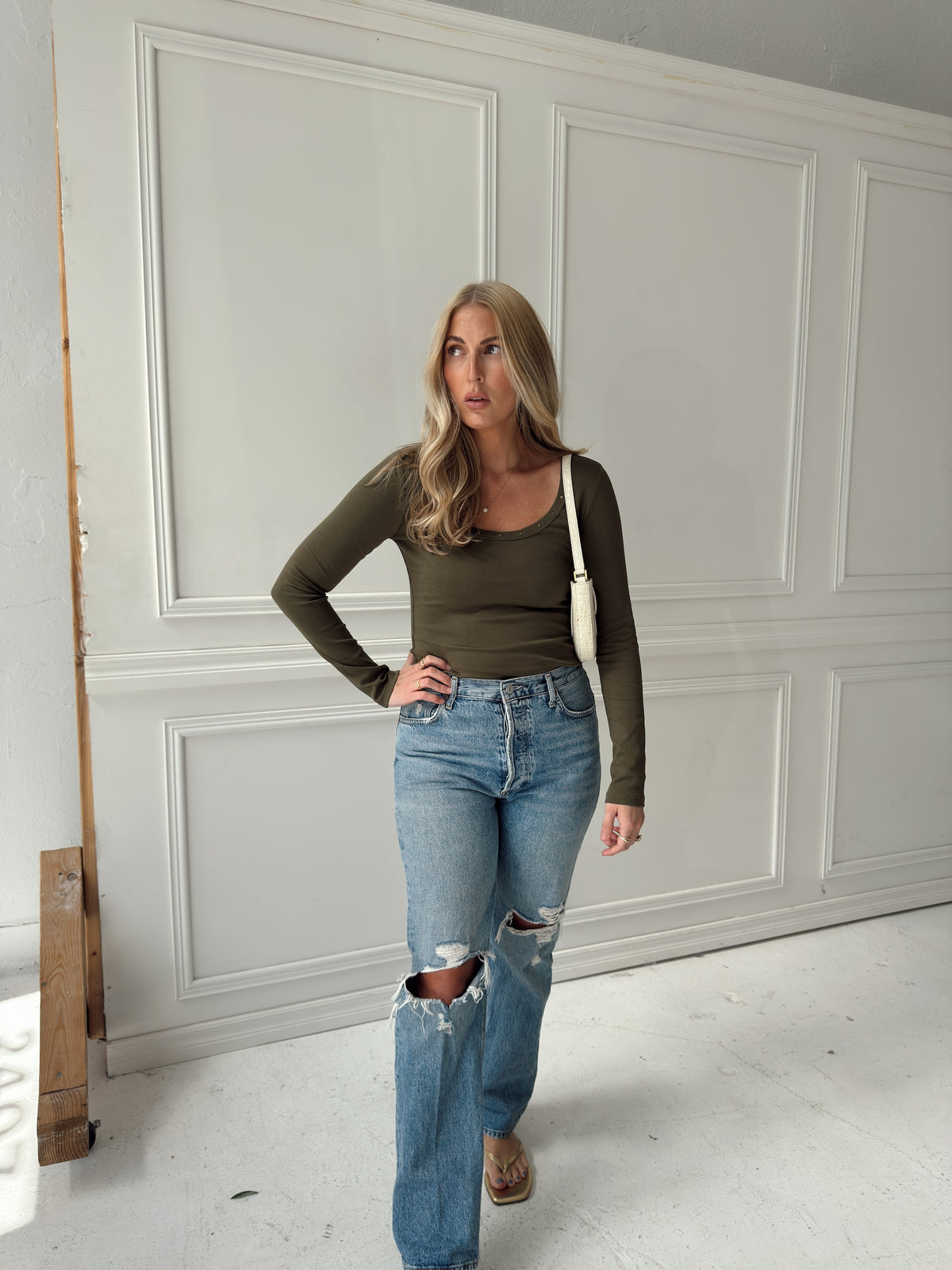 A woman stands in a room with white paneled walls wearing a fitted olive green long-sleeve top, distressed light-wash blue jeans, and brown flat shoes, with a white shoulder bag.