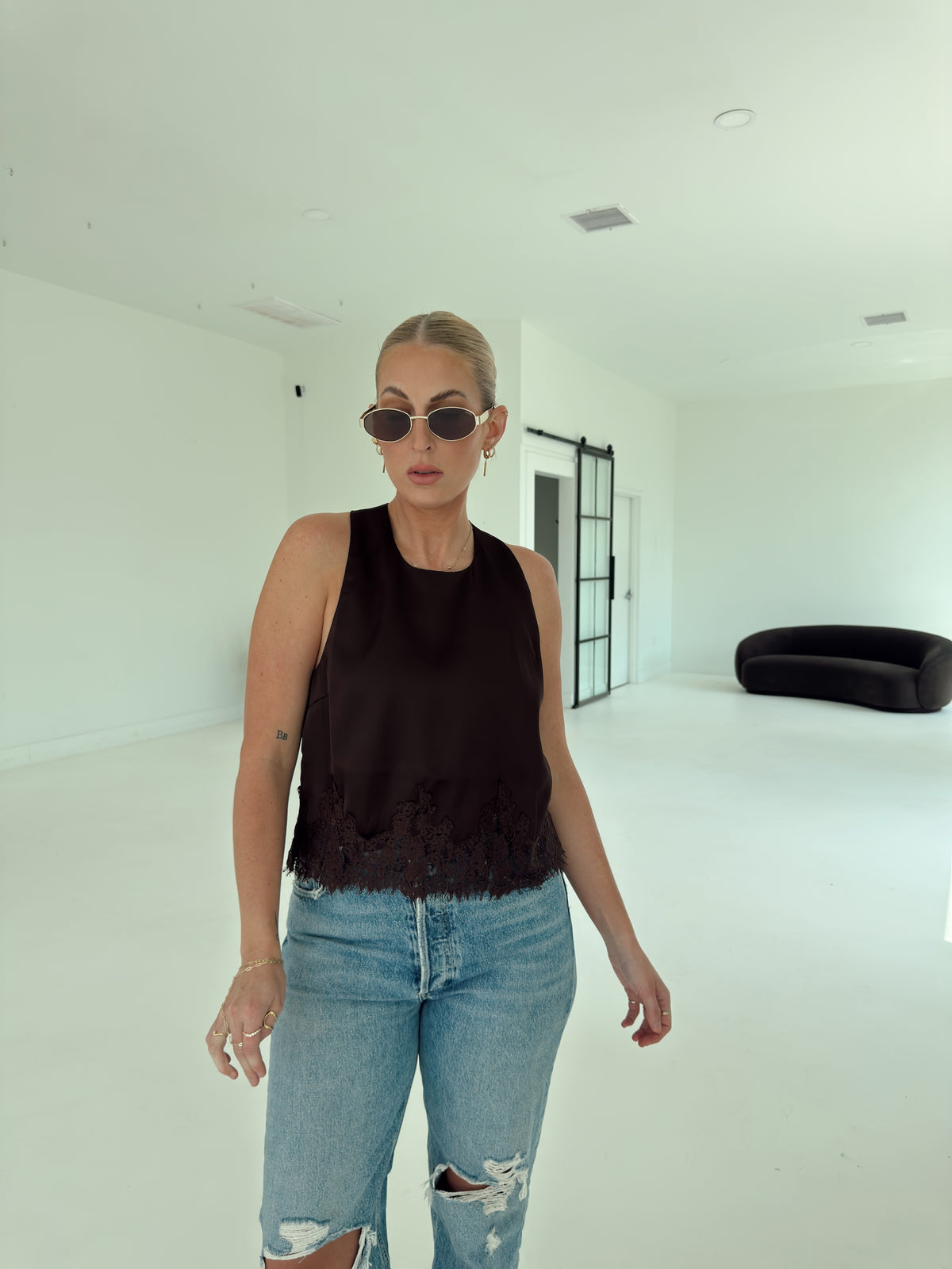A woman posing in a minimalist white studio setting, wearing a brown sleeveless top, light-wash distressed blue jeans, and white pointed-toe heels.