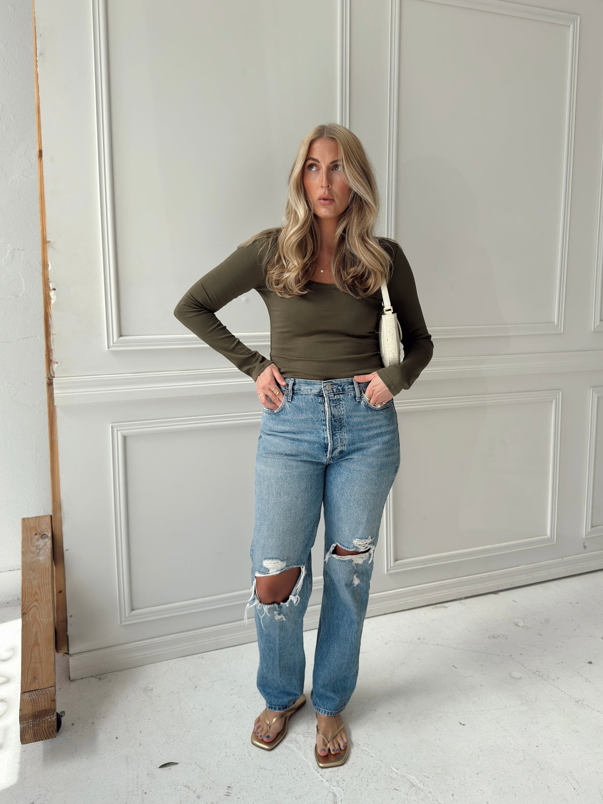 A woman posing in a room with white paneled walls, wearing an olive green long-sleeve top, light-wash distressed blue jeans with ripped knees, and brown sandals, while carrying a white shoulder bag.