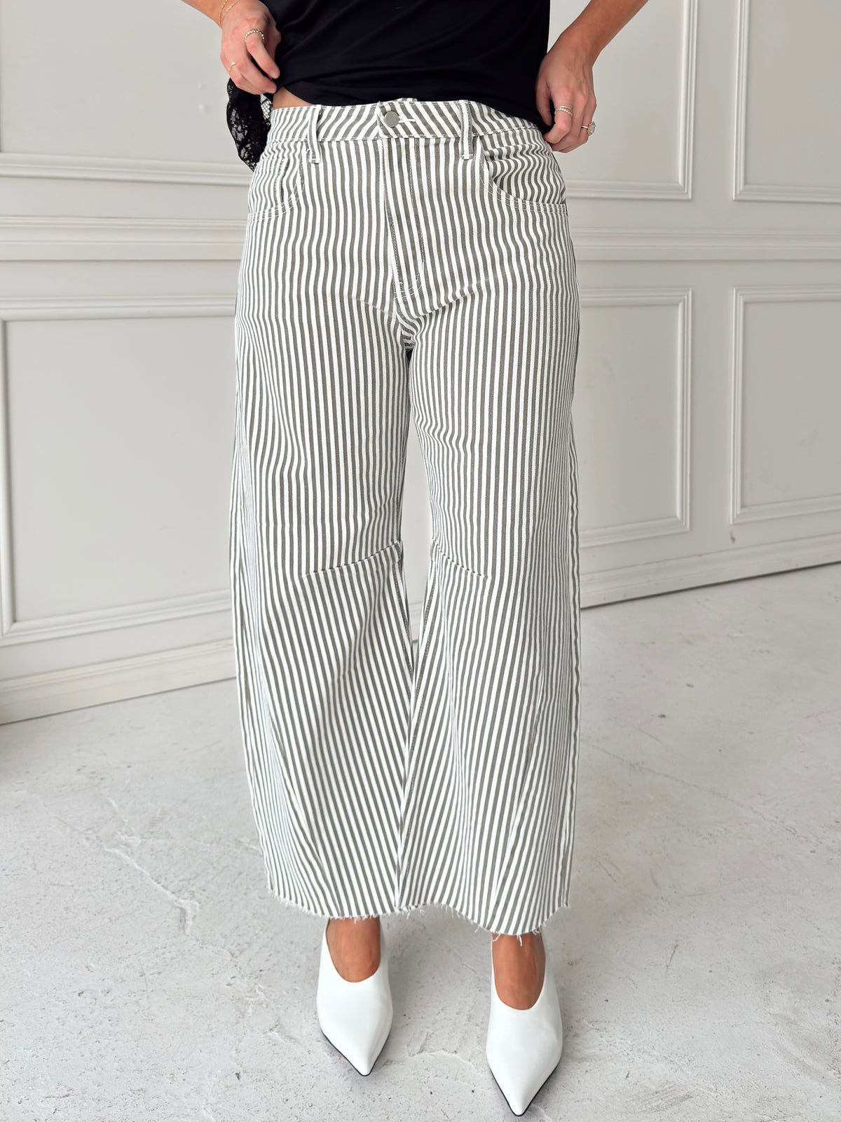 A woman modeling a pair of wide-leg barrel jeans with thin olive and white vertical stripes, paired with a black t-shirt,  and white pointed-toe heels, standing in front of a white paneled wall.