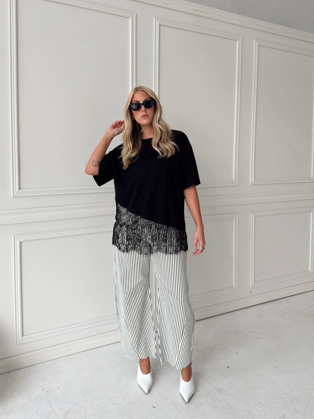 A woman modeling a black asymmetrical top with a sheer black lace hem, paired with white and olive vertically striped pants, standing in front of a white paneled studio backdrop.