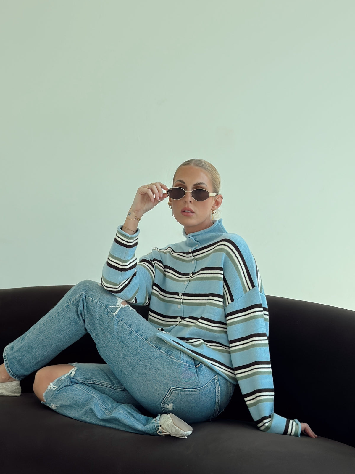 A woman sits on a dark couch wearing a light blue striped turtleneck sweater, distressed light-wash jeans, and sunglasses against a pale mint wall.