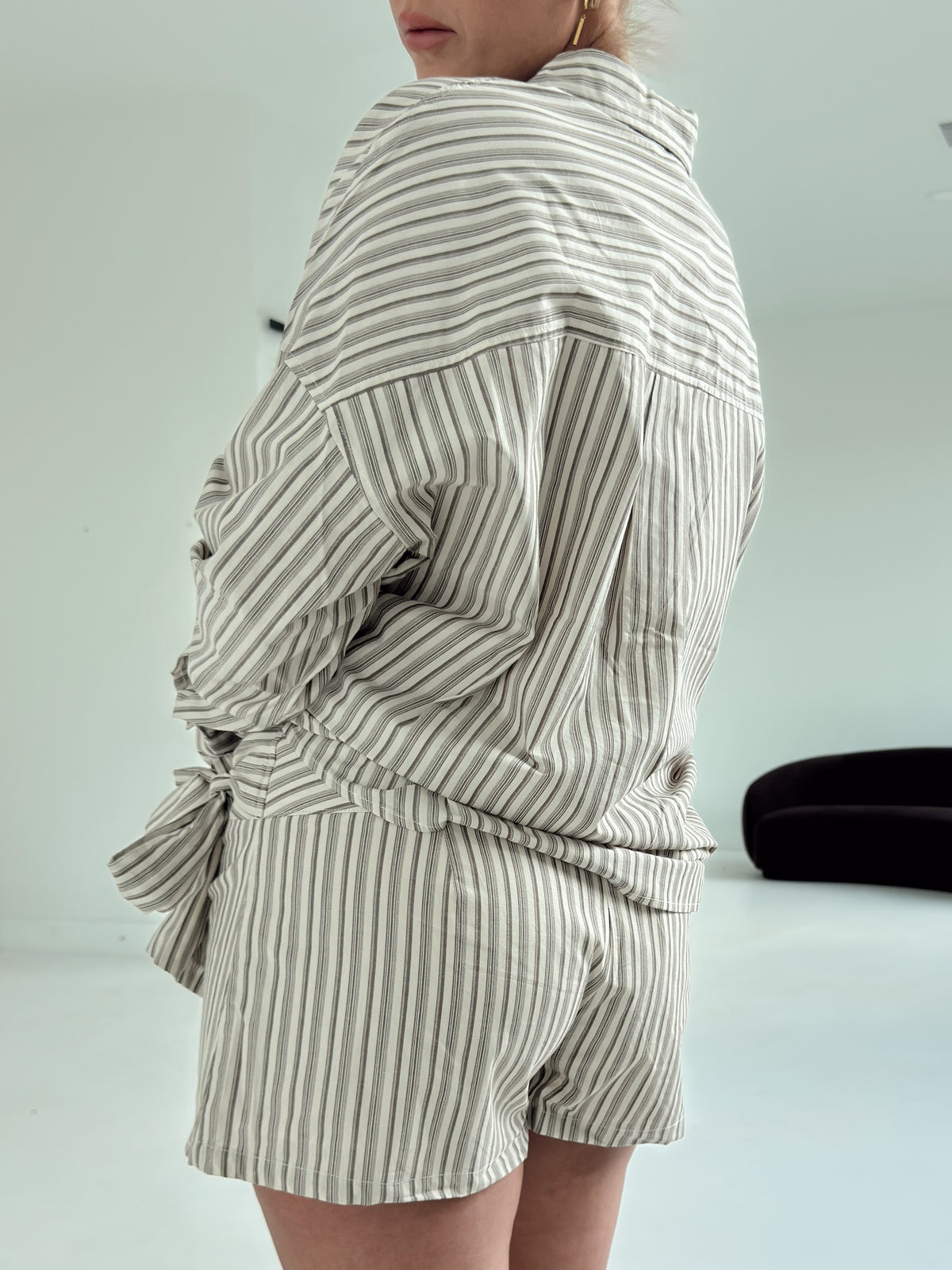 A model shown from the back wearing a matching two-piece set consisting of a long-sleeve button-up shirt and shorts, both featuring vertical stripes in beige and white.