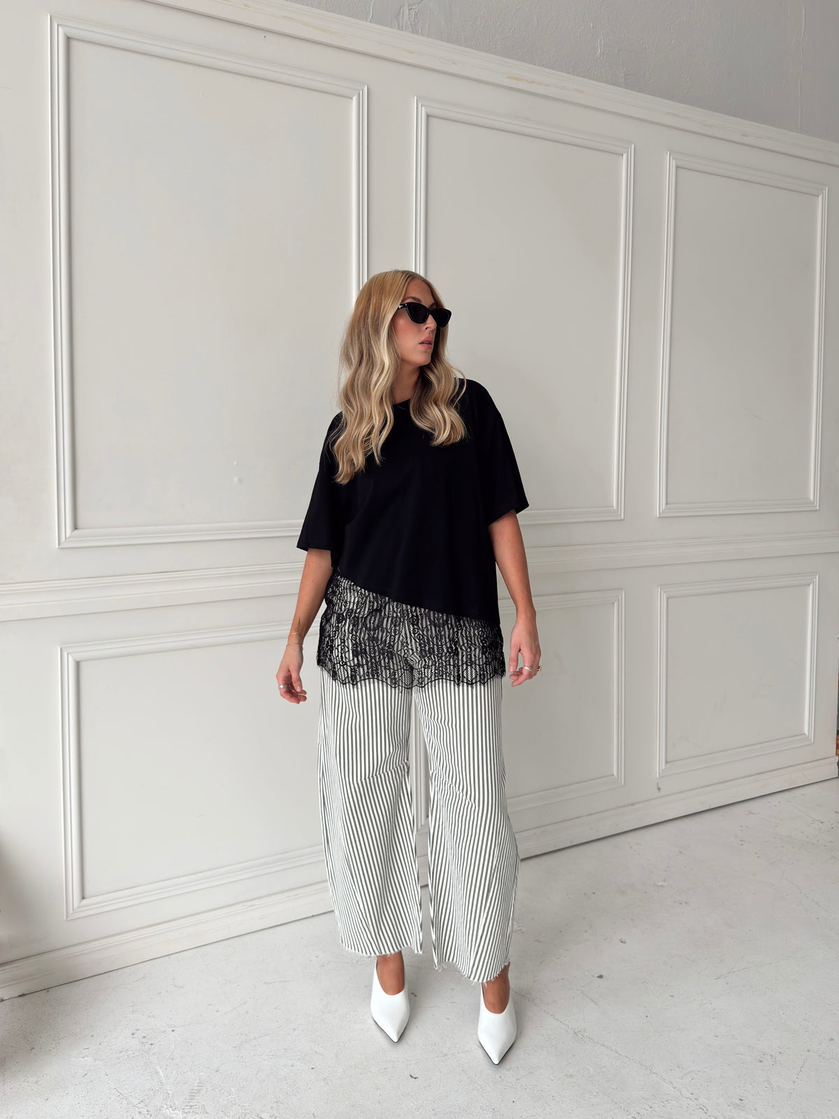 A woman modeling a pair of wide-leg barrel jeans with thin olive and white vertical stripes, paired with a black t-shirt, black sunglasses, and white pointed-toe heels, standing against a white paneled wall.
