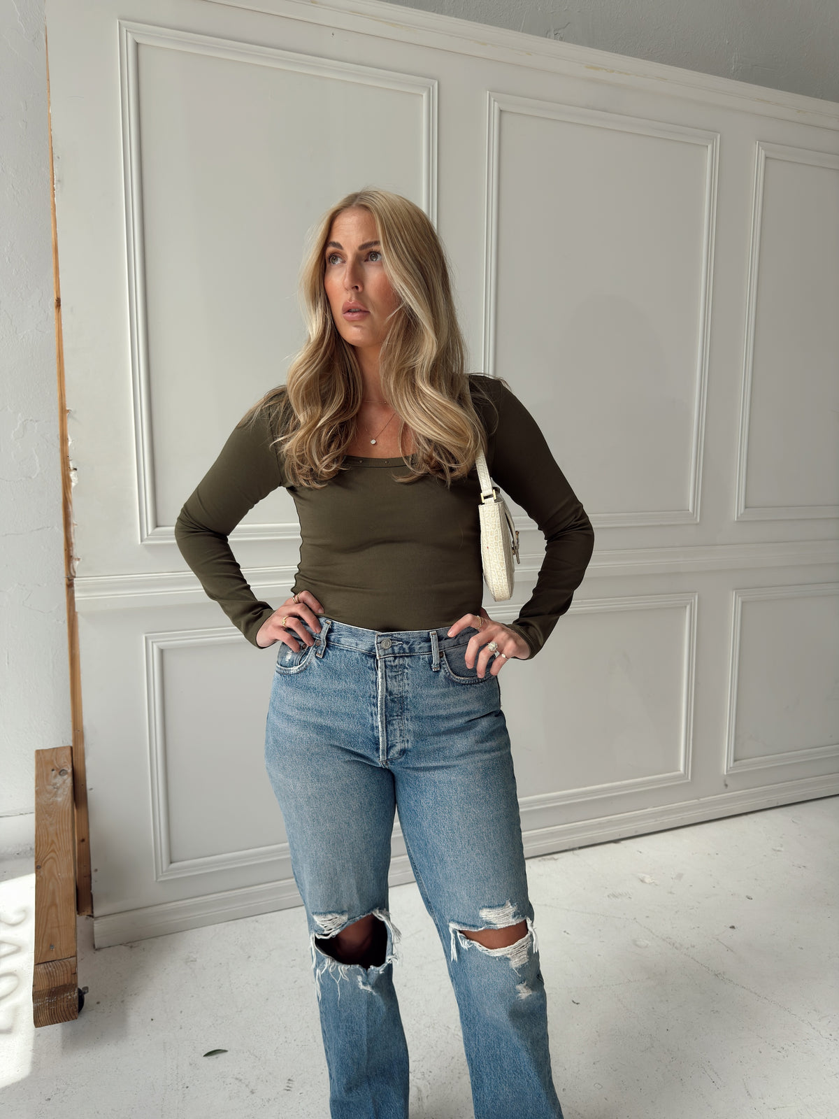 A woman posing in a room with white paneled walls, wearing an olive green long-sleeve scoop-neck top and light-wash distressed blue jeans.