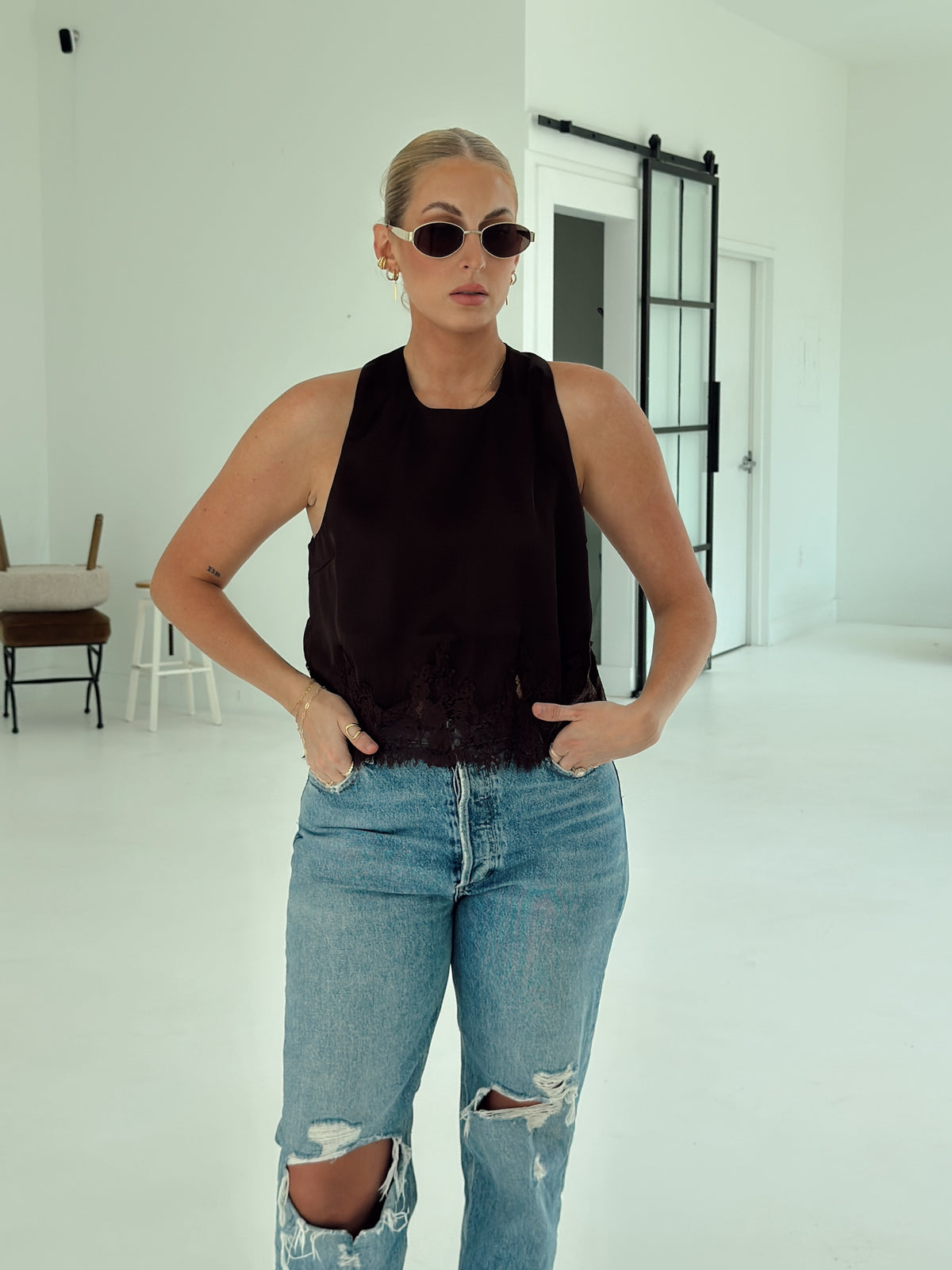 A woman modeling a sleeveless brown top with a textured, fringed hem and light-wash distressed blue jeans in a bright, white studio setting.