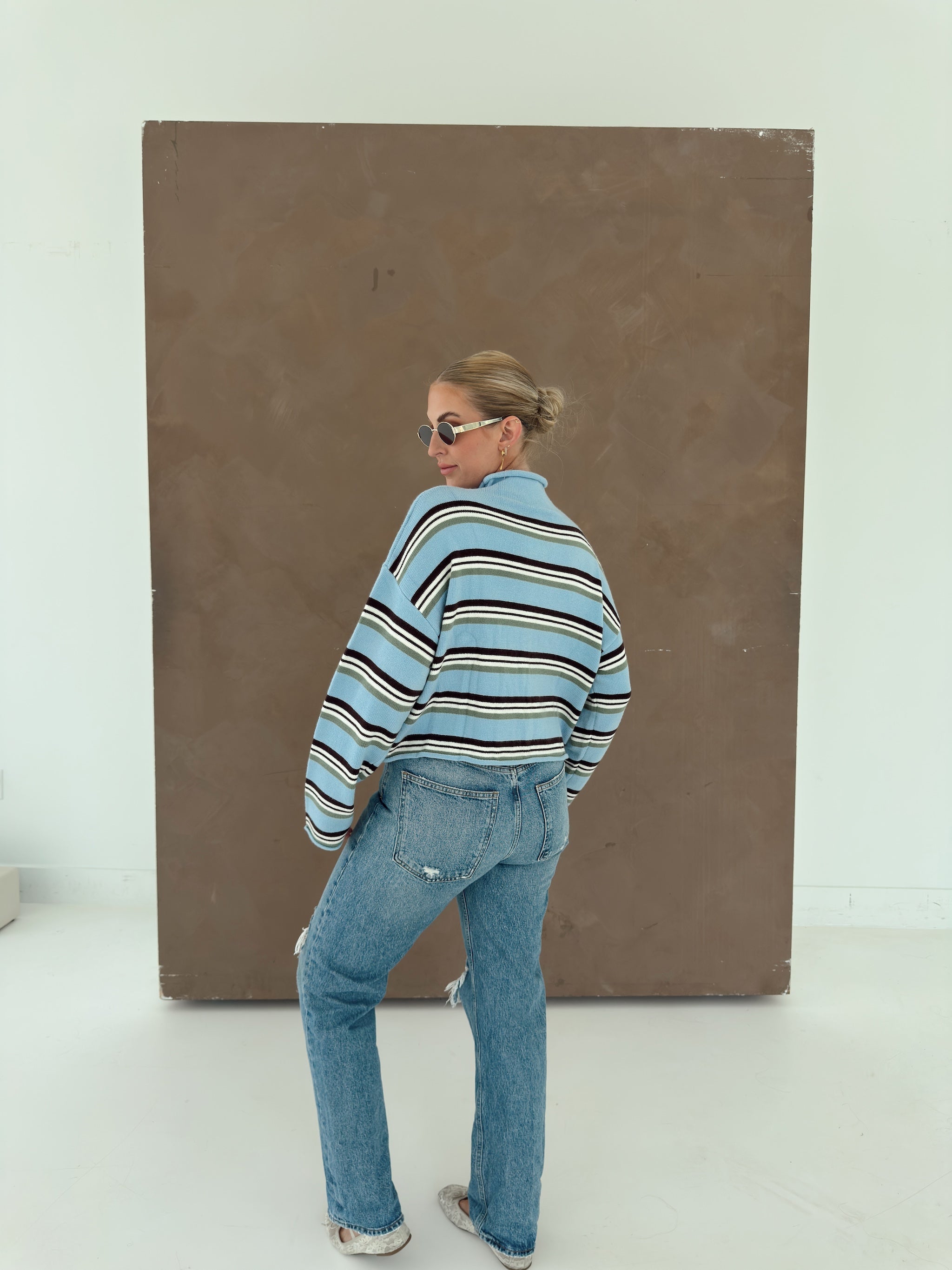 A woman wearing a light blue striped knit sweater, blue jeans, and sunglasses stands in profile in front of a large brown rectangular art piece on a white studio background.
