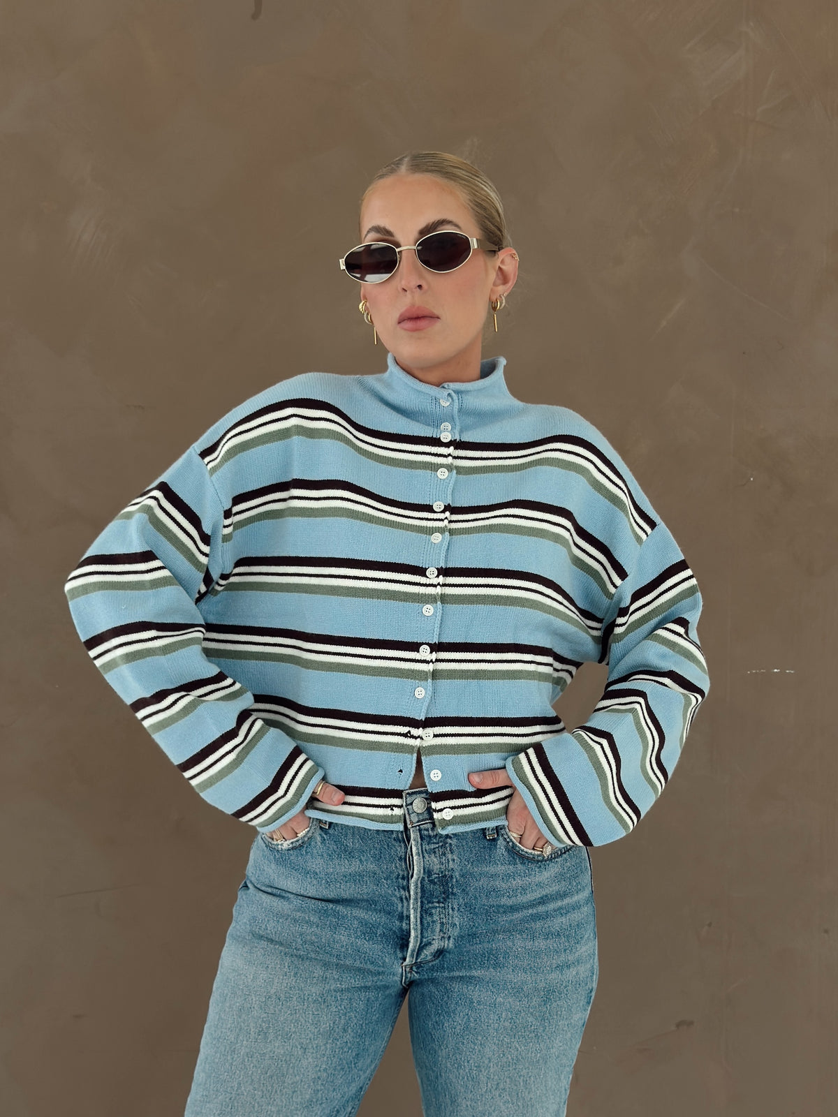 A person wearing a light blue knit cardigan with horizontal brown, white, and olive green stripes, paired with light-wash jeans and lace flats.