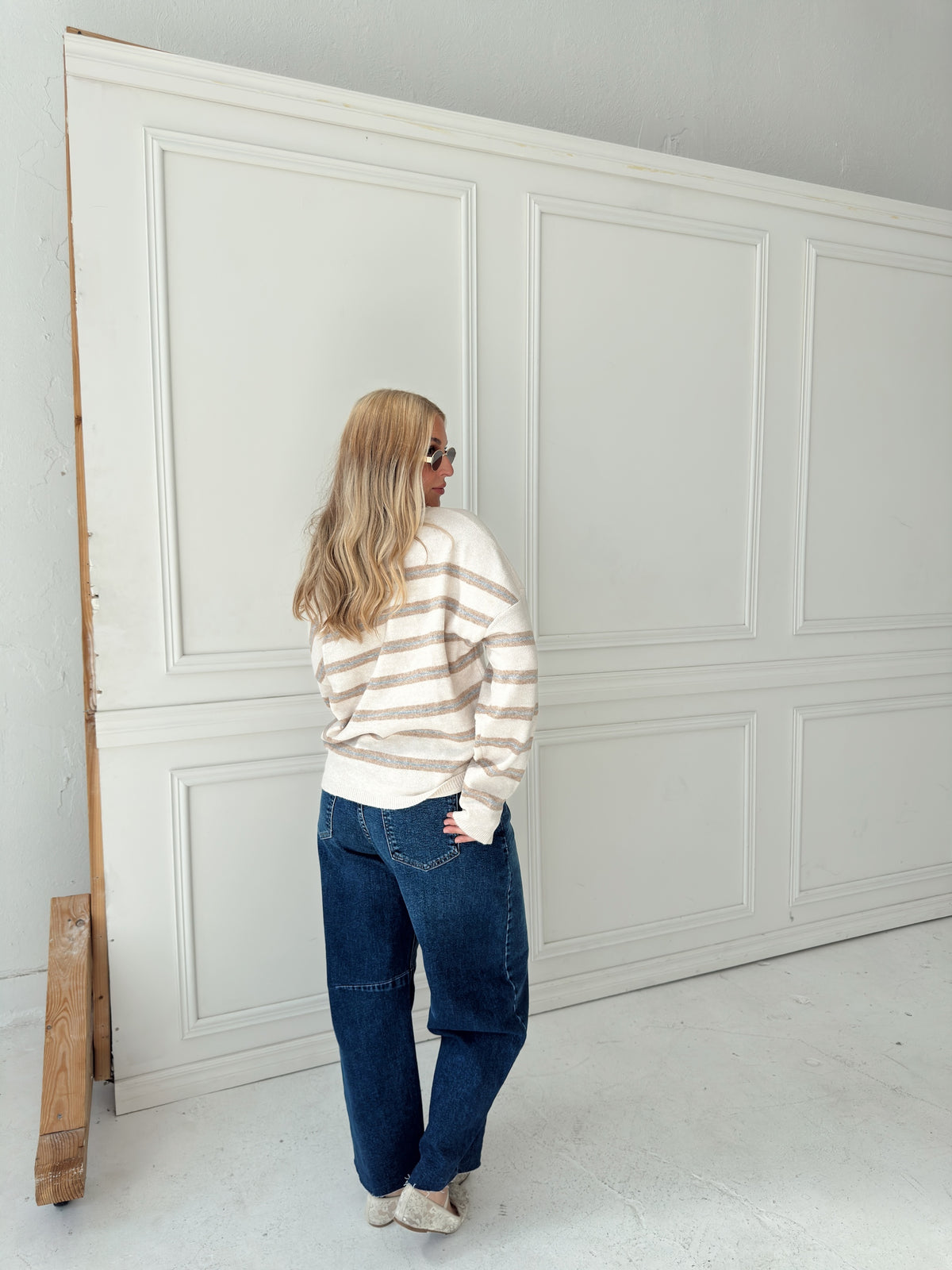 A woman stands with her back to the camera wearing a cream, light blue, and beige horizontally striped knit sweater and dark blue jeans, posing in front of a large white paneled wall.