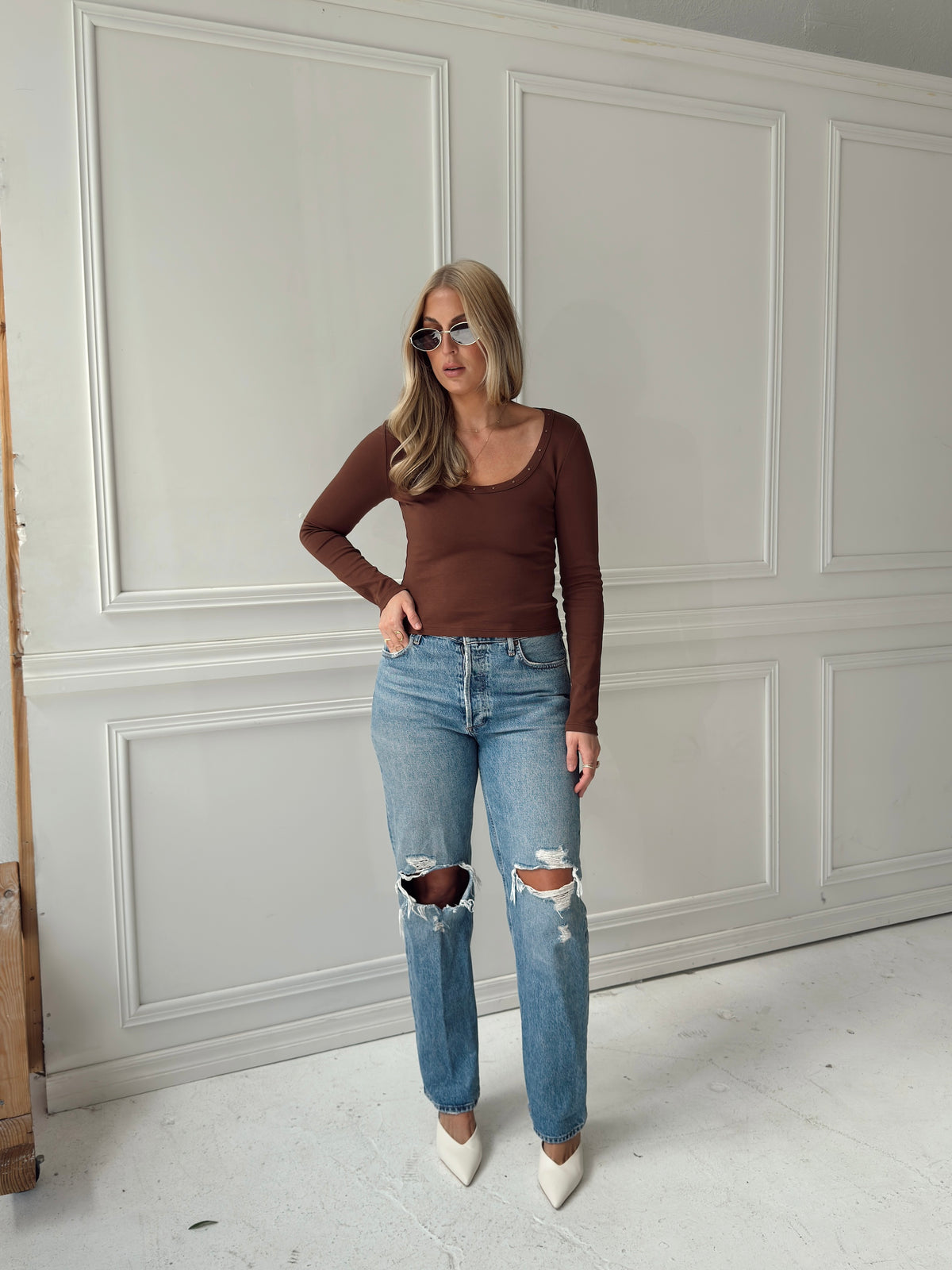 A woman stands in a room with white paneled walls wearing a brown long-sleeve scoop-neck top, light-wash distressed blue jeans with ripped knees, and white pointed-toe heels.