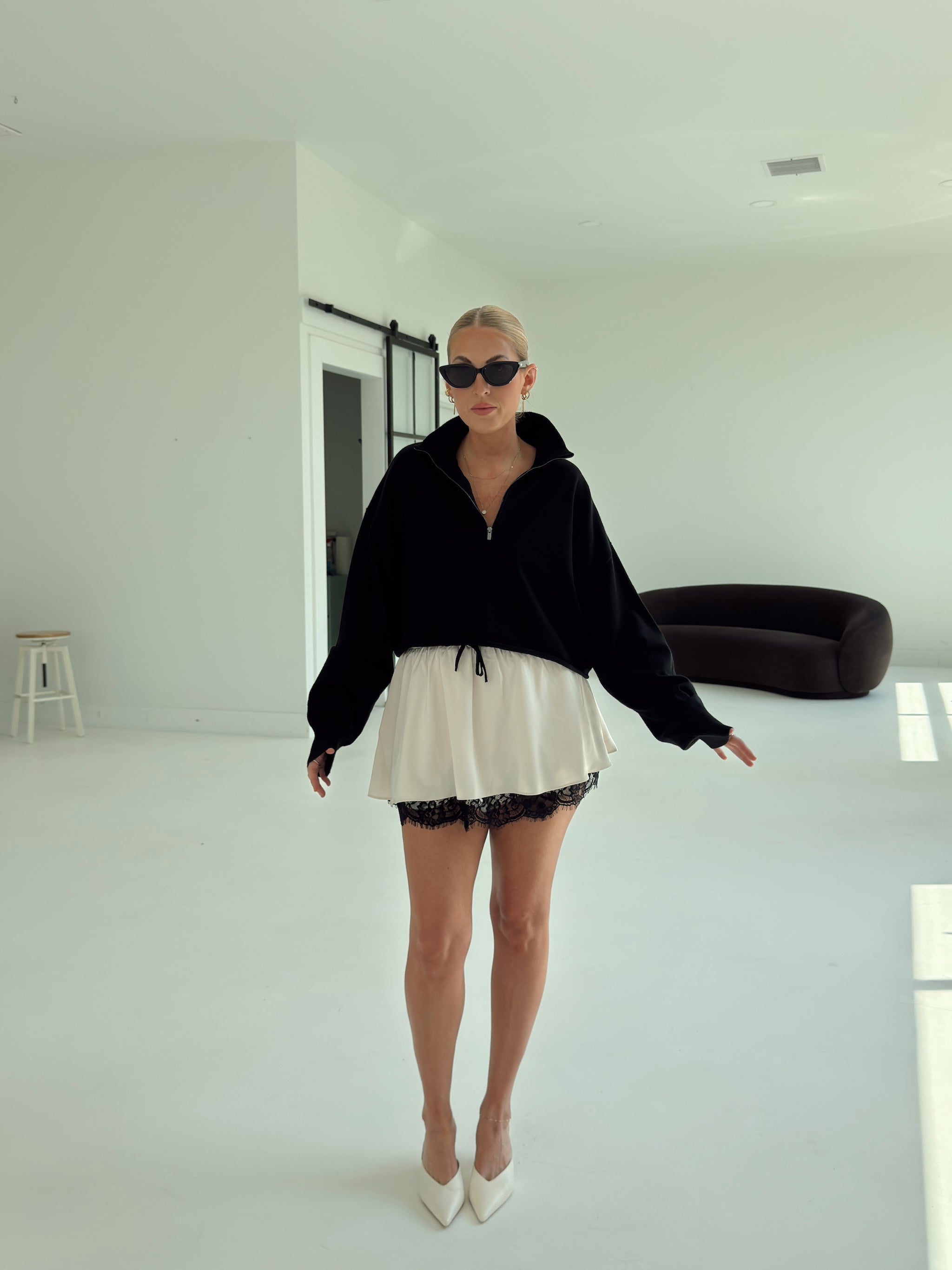 A woman modeling a black zip-up hoodie and a white ruffled mini skirt with black lace trim, styled with white pointed-toe heels and black sunglasses in a bright, minimalist white room.