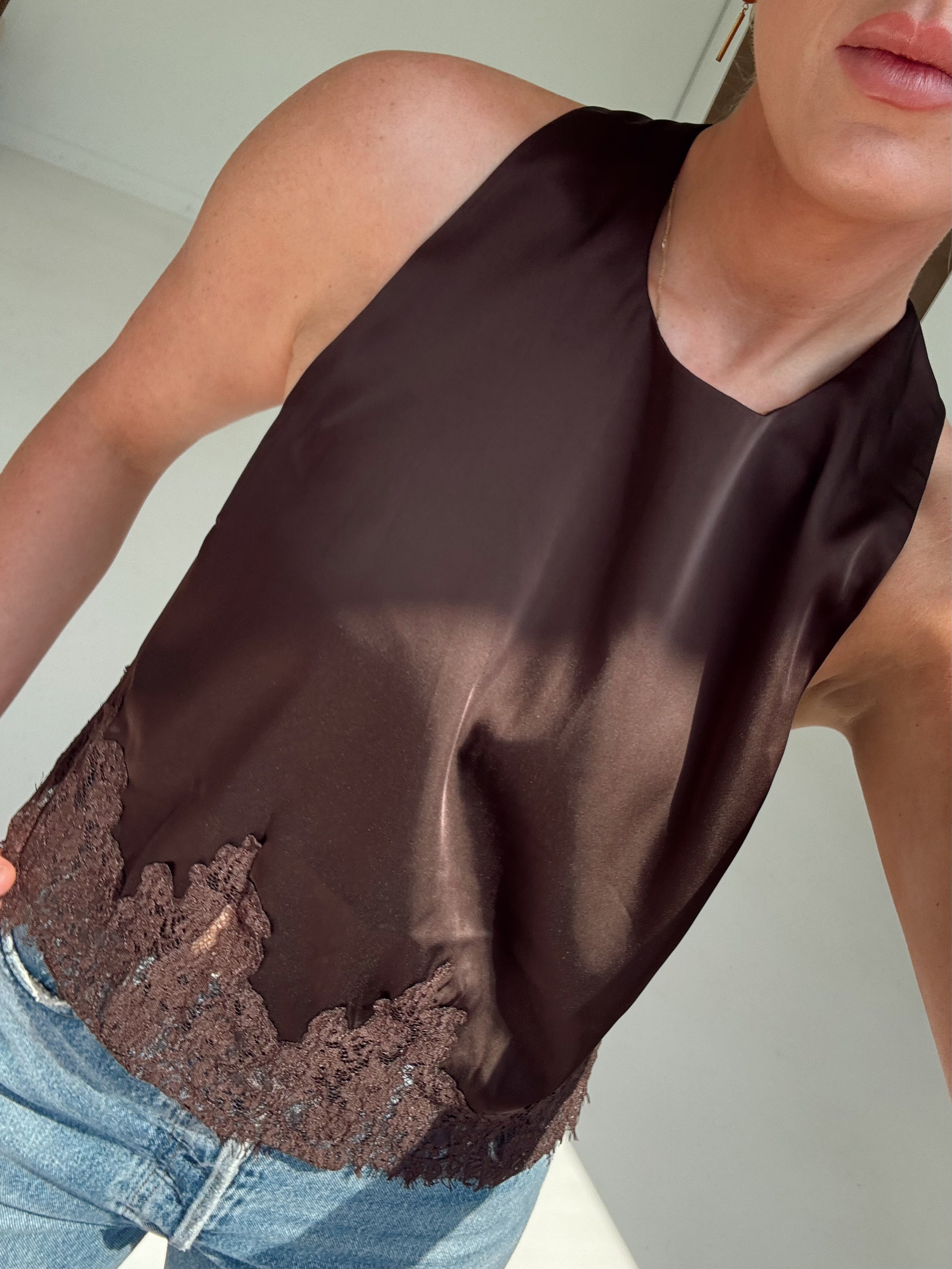 A close-up of a person wearing a sleeveless dark brown satin top with a lace-trimmed hem, paired with light blue jeans.