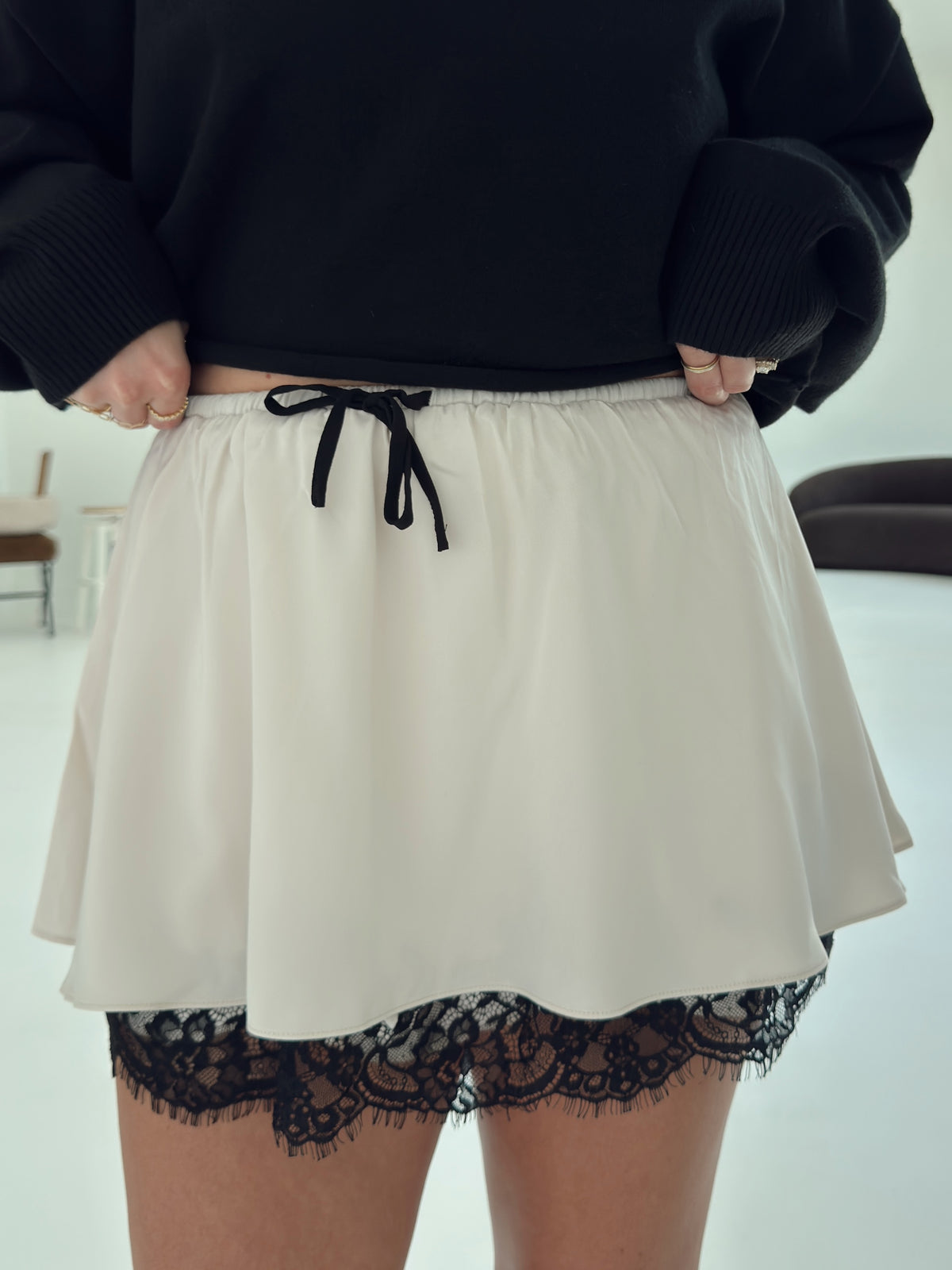 A person wearing a short cream skirt with a black drawstring and black lace trim at the hem, paired with a black sweater. The photo is cropped from the chest down in an indoor setting.