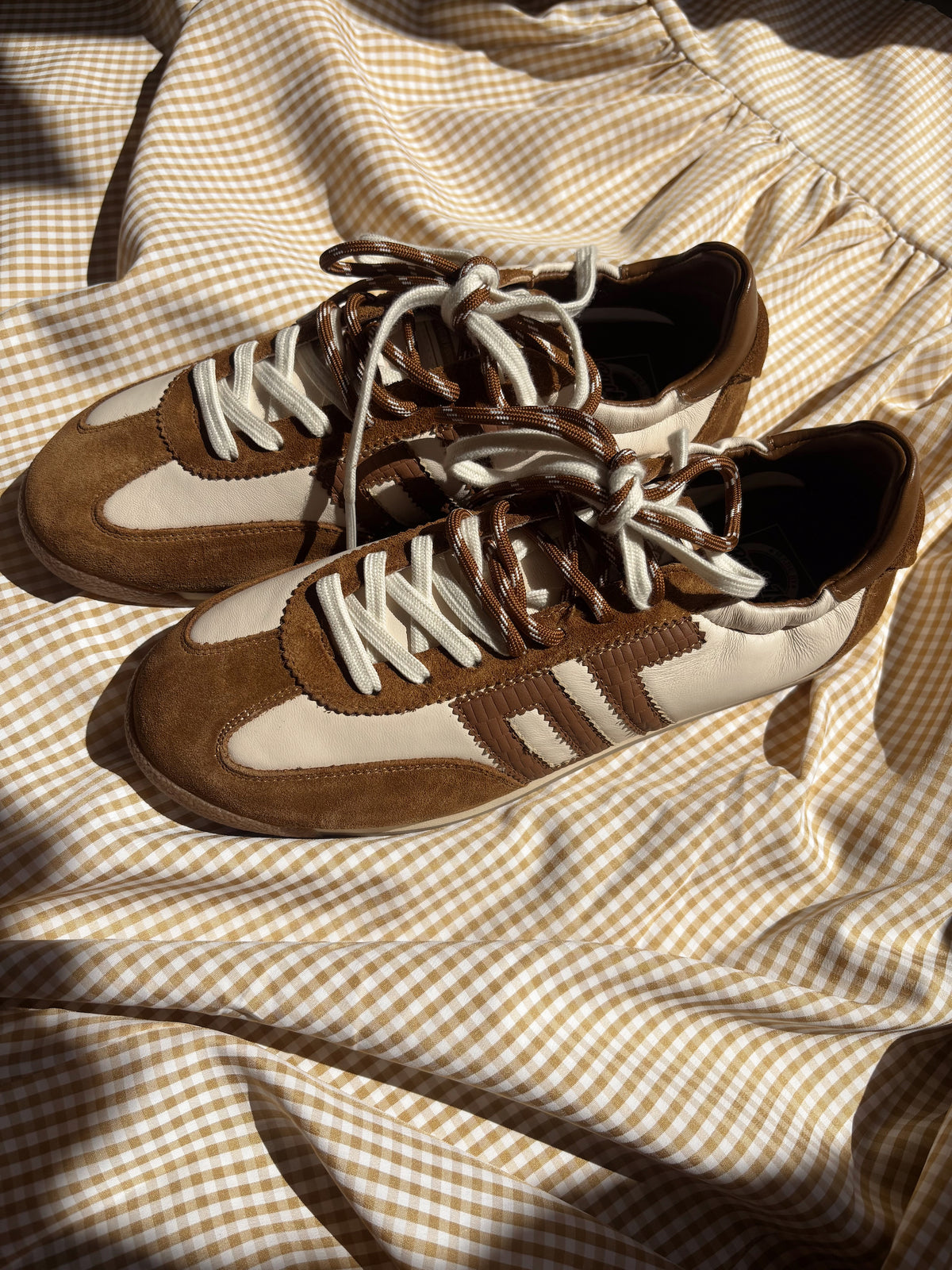 A pair of brown and cream suede sneakers with white laces, arranged on a brown and cream gingham checkered fabric under warm, direct sunlight.